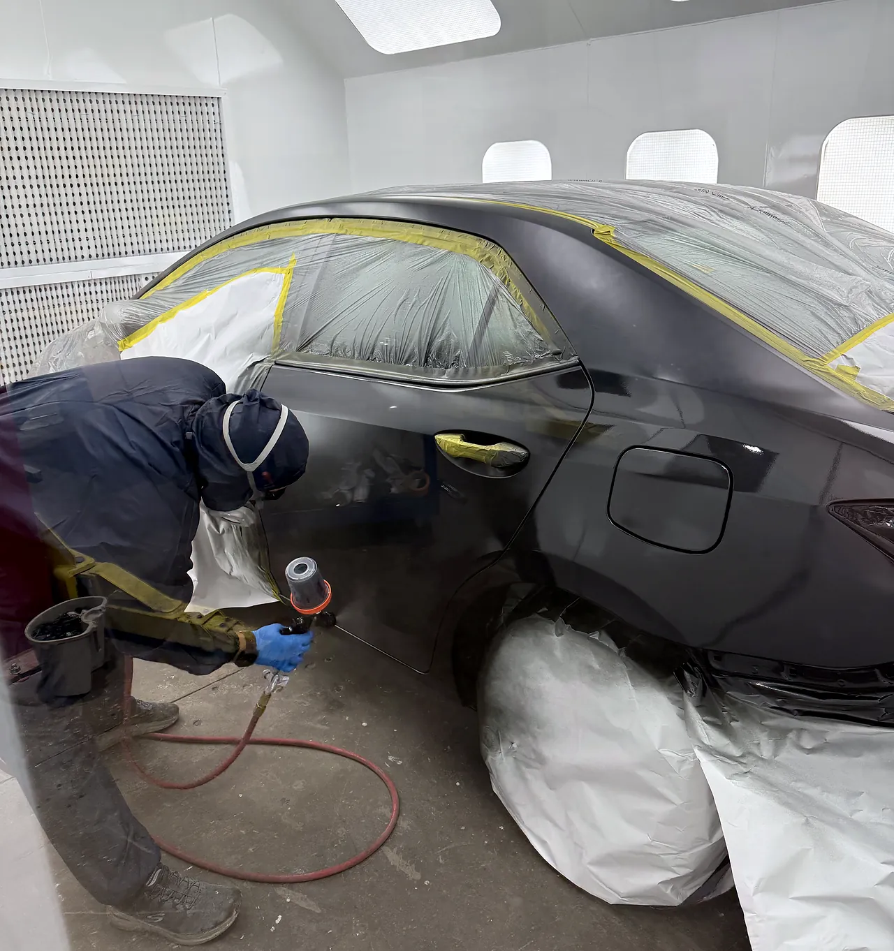 Sultan Motors refinishing technician in a full spray suit and respirator laying base coat inside the paint booth