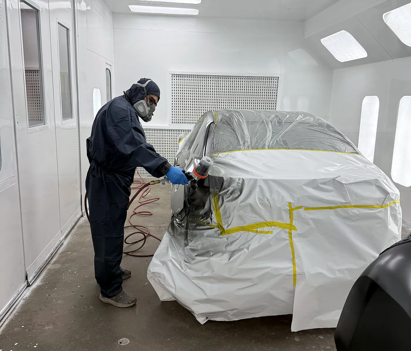 Technician in full protective equipment applying base coat to a masked vehicle in the Sultan Motors paint booth